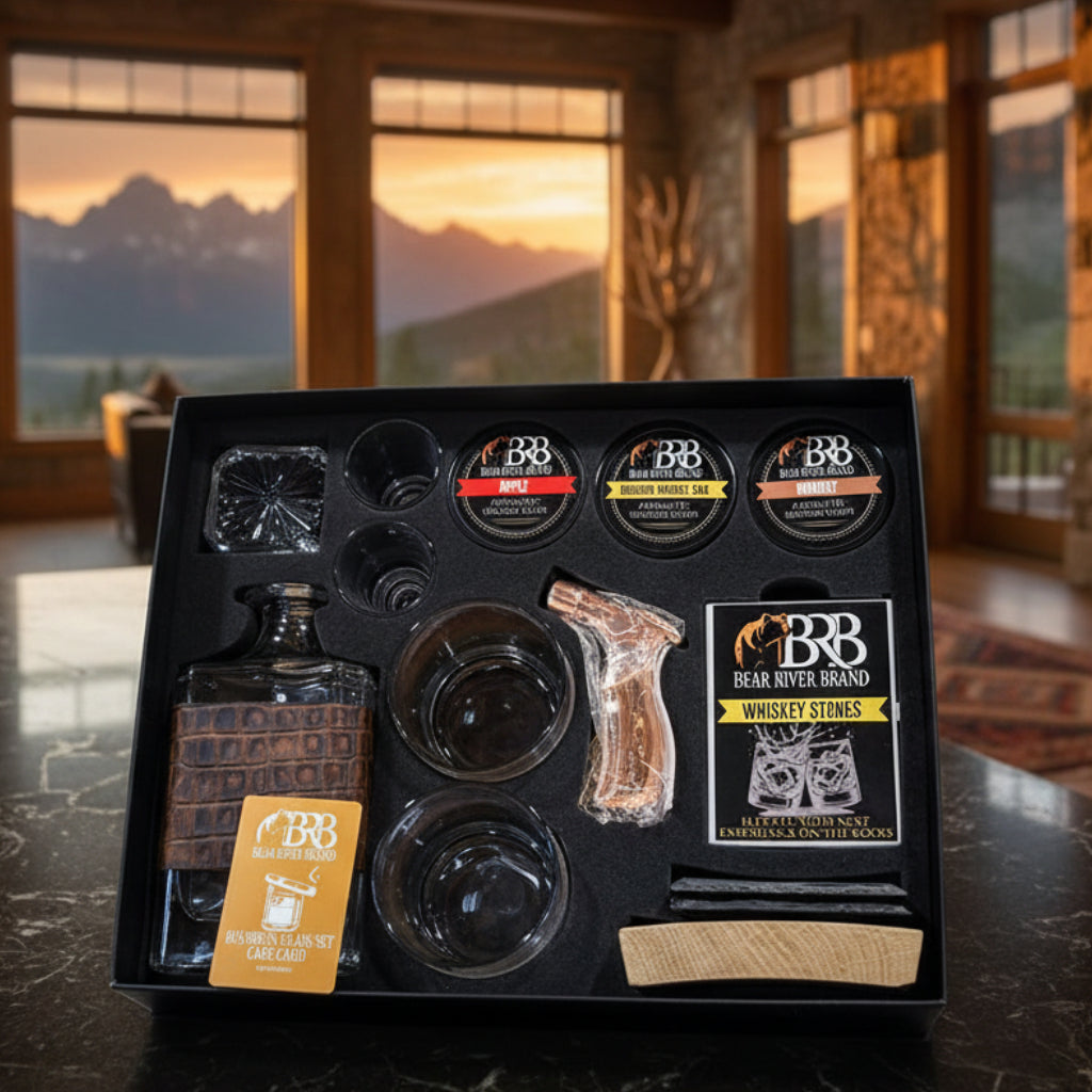 Bear River Brand whiskey gift set displayed in a black presentation box on a dark marble surface, with a scenic mountain landscape visible through large windows in the background. The set includes a whiskey decanter, 2 rock glasses, 2 shot glasses, 3 smoke flavors, a torch, whiskey stones, slate coasters, whiskey barrel cocktail smoker, and a branded care card.